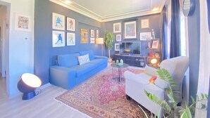 Een woonkamer met een blauwe bank, een witte fauteuil, een televisie en verschillende ingelijste foto's aan de muur.