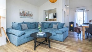 Egy modern nappali, melynek berendezésében szerepel egy kék színű, moduláris kanapé, egy kerek dohányasztal, valamint egy étkezősarok, ahol egy fa asztal és székek találhatók.