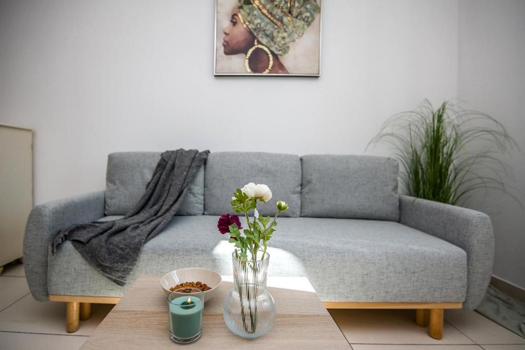 A modern living room with a grey sofa, a small table with a vase of flowers, a bowl of nuts, and a candle.