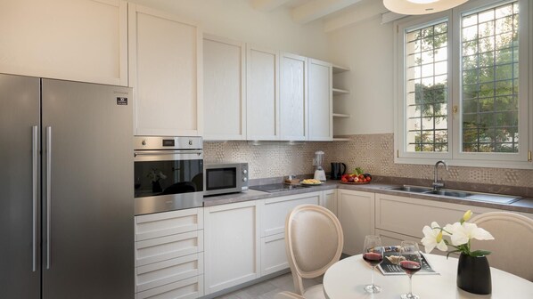 A modern kitchen with stainless steel appliances, white cabinetry, a dining area with a table and chairs, and a window with a view of trees. - Villa Pergola 8 in Bologna (Castel San Pietro Terme)