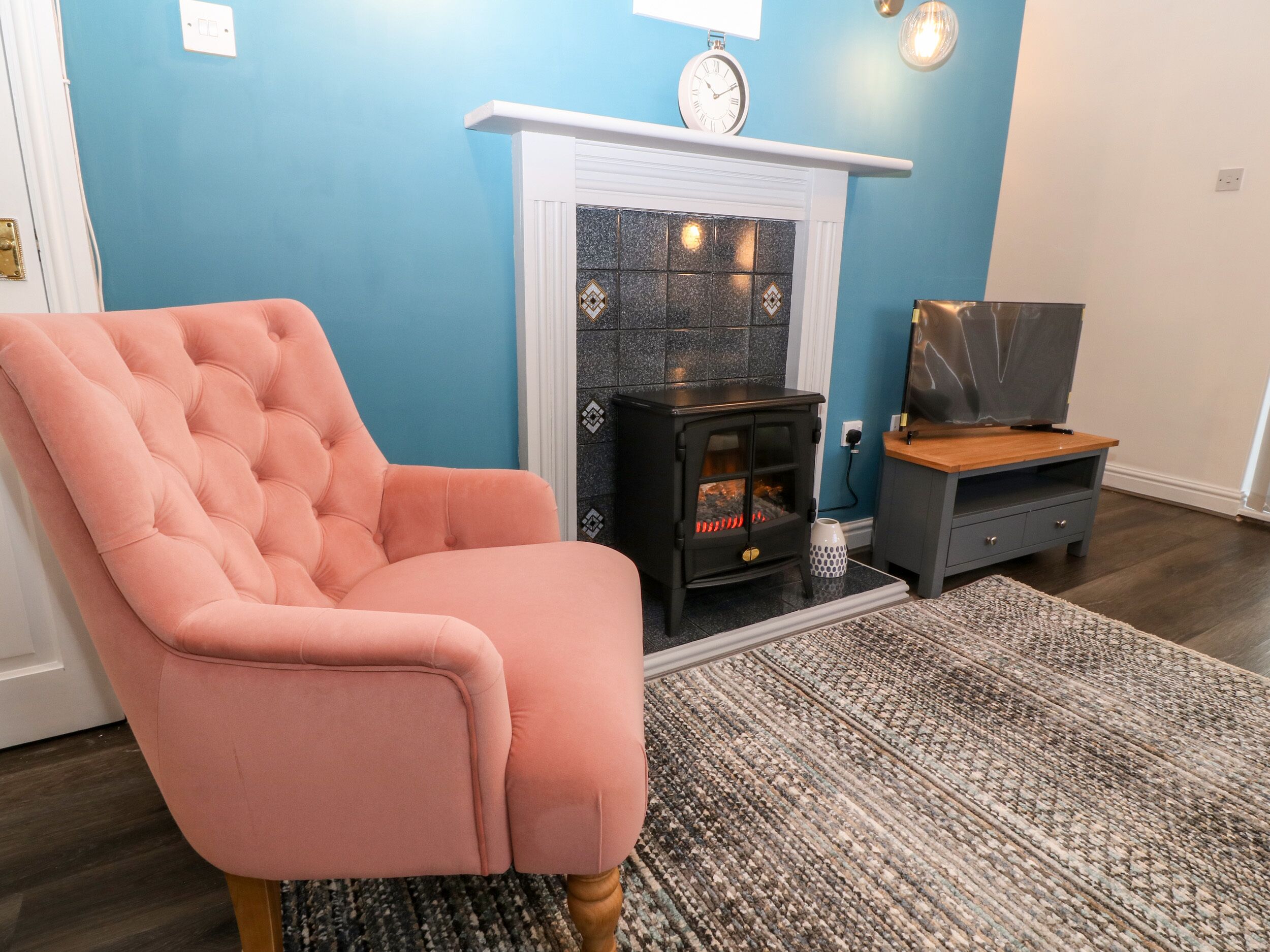 Un salon avec un fauteuil rose à capitons, un poêle à bois noir, un téléviseur posé sur un meuble en bois et un tapis à motifs.