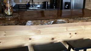 Une cuisine avec des meubles en bois, un réfrigérateur en acier inoxydable, une machine à café et un bocal en verre rempli de fruits. Il y a trois tabourets noirs devant le comptoir.