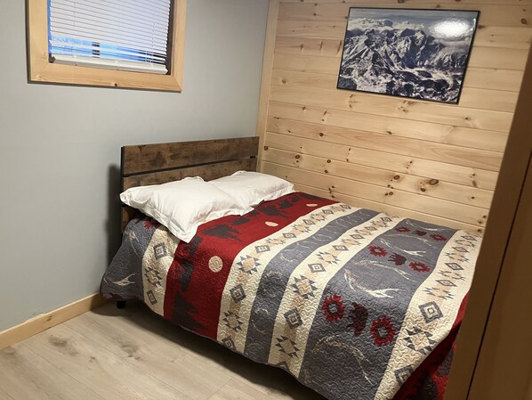 A single bed with a patterned bedspread, a white pillow, and a wooden headboard in a room with wooden panel walls and a framed picture above the bed.