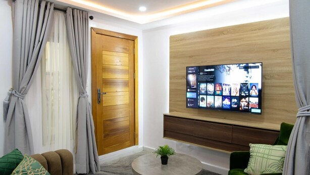 Un salon moderne avec une porte en bois, un téléviseur fixé au mur diffusant un service de streaming, une table basse ronde ornée d’une plante et des canapés recouverts d’un tissu vert.