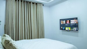 Une chambre d’hôtel avec un lit, des rideaux beiges, un téléviseur fixé au mur diffusant du contenu YouTube et un mur blanc.