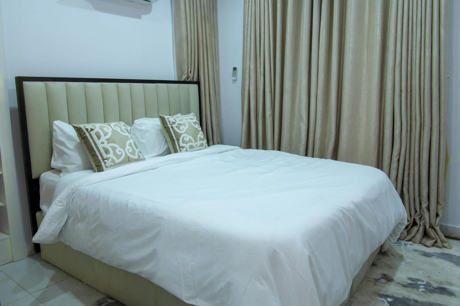 A neatly made bed with white bedding and patterned pillows, a beige headboard, and a curtain in the background.