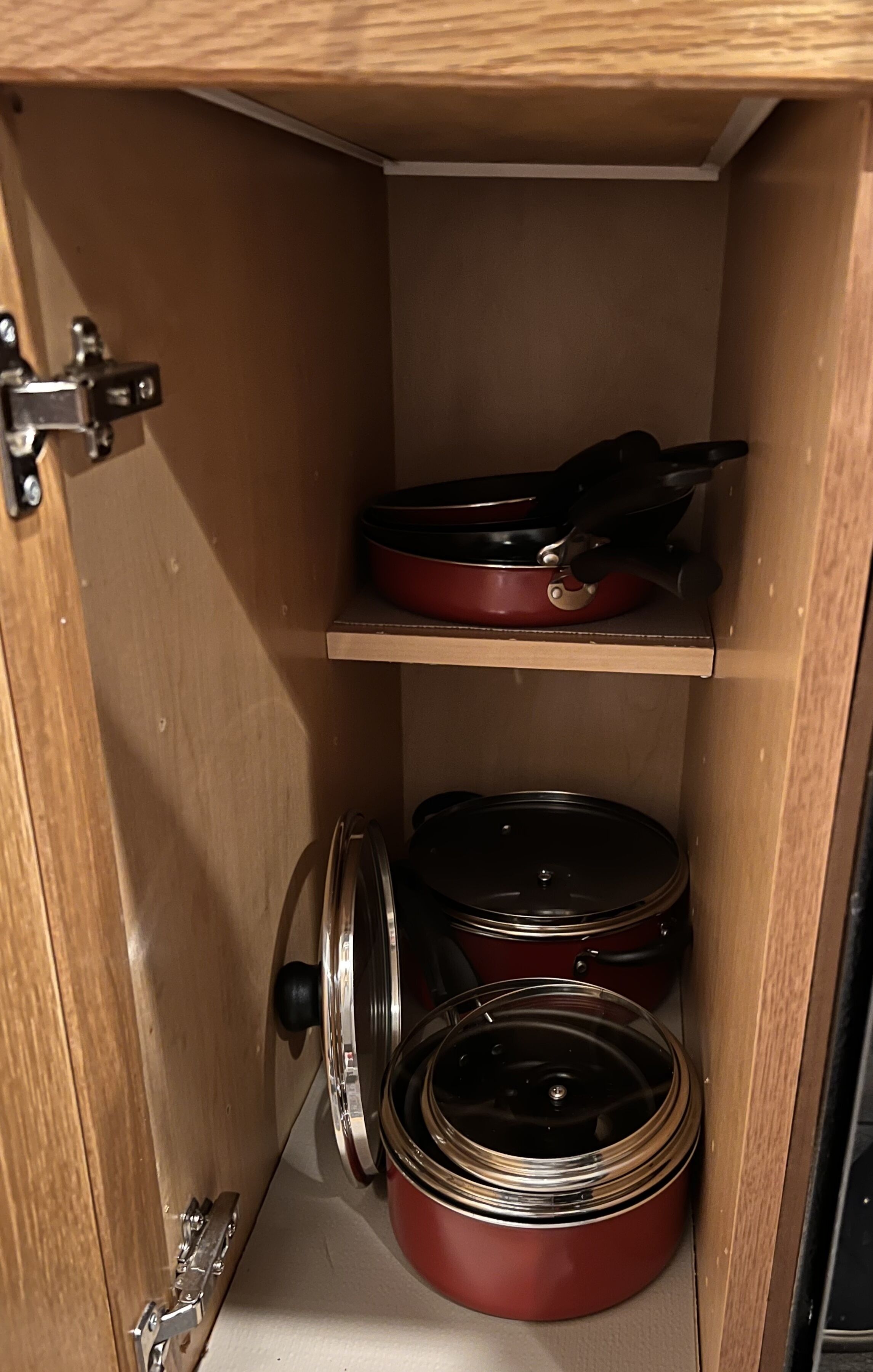 A kitchen cabinet with a red frying pan on the top shelf and two red pots with lids on the bottom shelf.