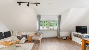 Eine moderne Lofts-Wohnung mit hohen Decken, einer gemütlichen Sitzecke mit Sofa und Sesseln, einem Fernseher auf einem weißen Schrank und einem großen Fenster mit Vorhängen.