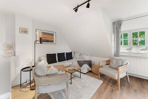Un loft moderno con techo inclinado, sofá blanco, sillón de mimbre, mesa de centro y vistas a la vegetación a través de una ventana.