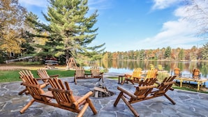 Een terras van natuursteen met houten Adirondack stoelen en een vuurkuil, met uitzicht op een meer met herfstbomen en een steiger.