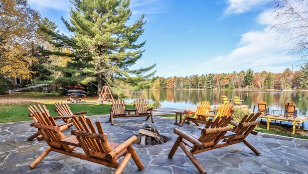 Une terrasse en pierre avec des fauteuils Adirondack en bois et un foyer, donnant sur un lac bordé d’arbres aux couleurs automnales et sur un quai.