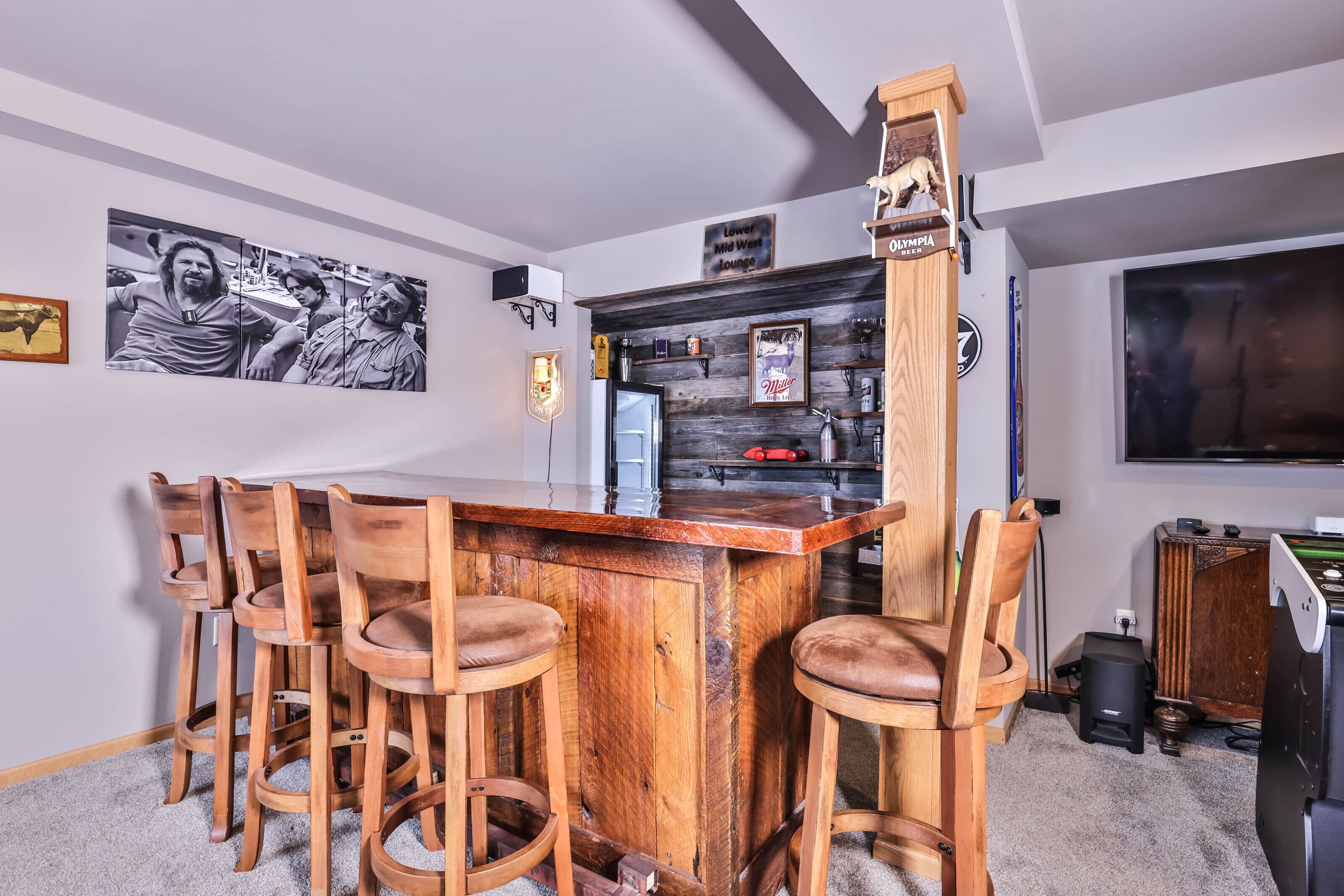 Uma área de bar com balcão de madeira e quatro banquetas, geladeira e prateleira com garrafas e um letreiro.