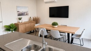 Una cocina moderna con un lavamanos doble, una mesa de comedor con sillas, un televisor montado en la pared y un mueble pequeño con objetos decorativos.