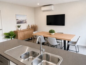 A modern kitchen with a double sink, a dining table with chairs, a TV mounted on the wall, and a small cabinet with decorative items.
