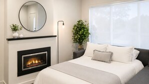 Un dormitorio con chimenea, una cama, una mesita de noche con una planta, un espejo y una ventana.