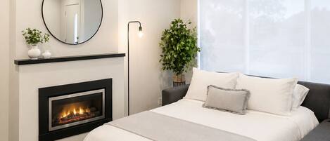 Un dormitorio con chimenea, una cama, una mesita de noche con una planta, un espejo y una ventana.