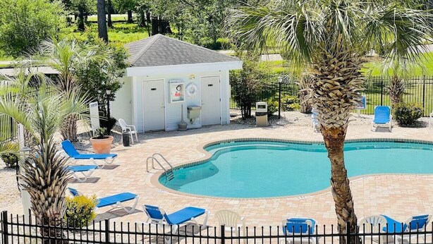 Una piscina con sillones azules, un edificio pequeño con puerta y ventanas, y follaje circundante.