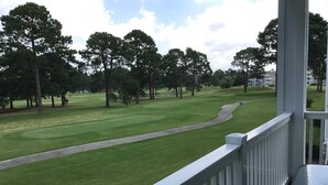 Vista de un campo de golf desde un balcón con barandilla, árboles y cielo despejado.