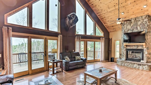 A spacious living room with a fireplace, large windows, and a stone accent wall. - Spacious Lead Getaway w/ Private Hot Tub & Deck! (Lead)