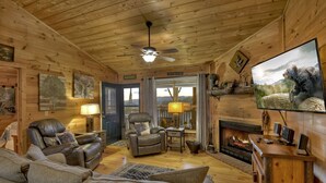 A cozy cabin interior with a leather sofa, a recliner, a fireplace, and a television on a wooden stand. - Overlook Oasis - Hot tub, firepit, and comfy king beds! (Blue Ridge)