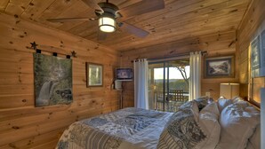 A wooden cabin bedroom with a bed, ceiling fan, artwork, and a television. - Overlook Oasis - Hot tub, firepit, and comfy king beds! (Blue Ridge)