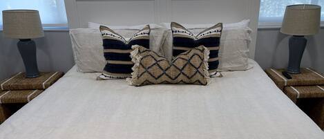 Una cama con sábanas blancas, dos mesitas de noche con lámparas y almohadones decorativos.