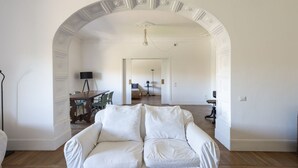 Un canapé blanc dans une pièce dotée d’une porte en arc donnant sur une autre pièce où se trouvent un lit et une lampe.