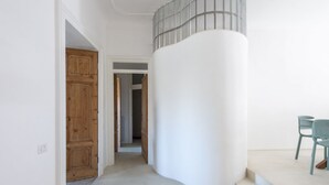 Un couloir avec une porte en bois, une colonne blanche et un escalier menant à un balcon où se trouvent des chaises.
