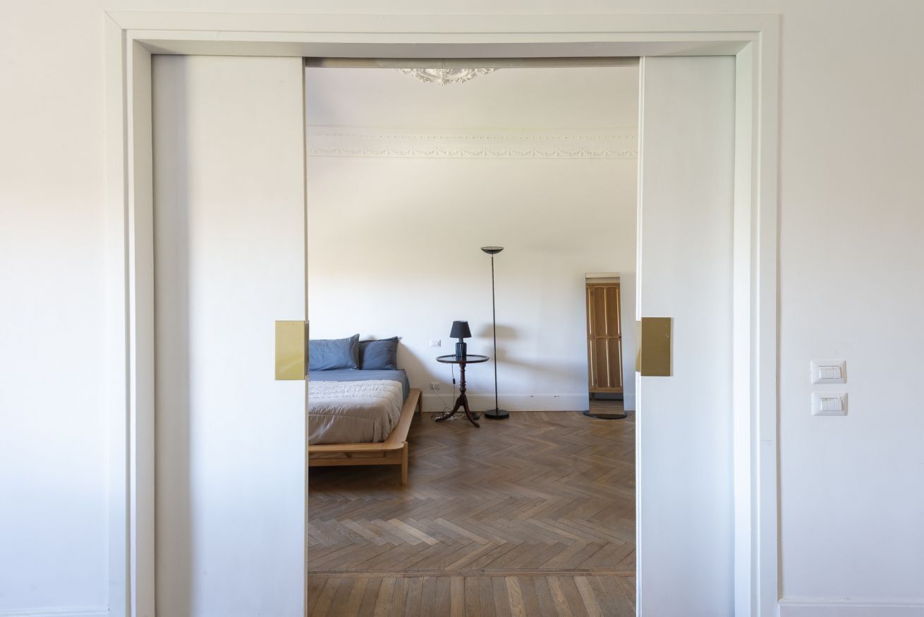 Une chambre à coucher avec un lit, une table de chevet, une lampe et une vue sur une autre pièce.
