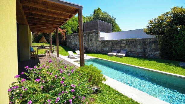 A modern outdoor pool area with a pergola, stone wall, and a small seating area with cushions.