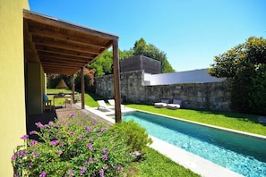 Un área de piscina moderna al aire libre con una pérgola, un muro de piedra y una pequeña zona de estar con cojines.