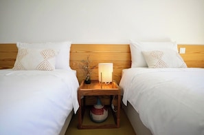 Habitación de hotel con dos camas, cada una con ropa blanca y almohadas decorativas. Hay una mesita de noche de madera entre las camas, con una lámpara y una cesta tejida.
