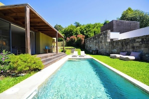 Un área de piscina moderna al aire libre con una piscina cristalina, una pérgola de madera y un muro de piedra.