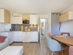 A compact kitchen with white cabinets, a coffee maker, and a toaster. There is a dining area with a wooden table and two chairs.