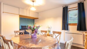 A dining area with a round table, chairs, a vase of flowers, and a view of the outdoors through a window with curtains. - Spacious apt 7 ppl, mountain view balcony (Saint-Sorlin-d´Arves)