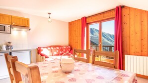 Ein gemütlicher Essbereich mit Tisch für zwei Personen, Holzstühle, eine rot gemusterte Couch und Bergblick durch große Fenster.