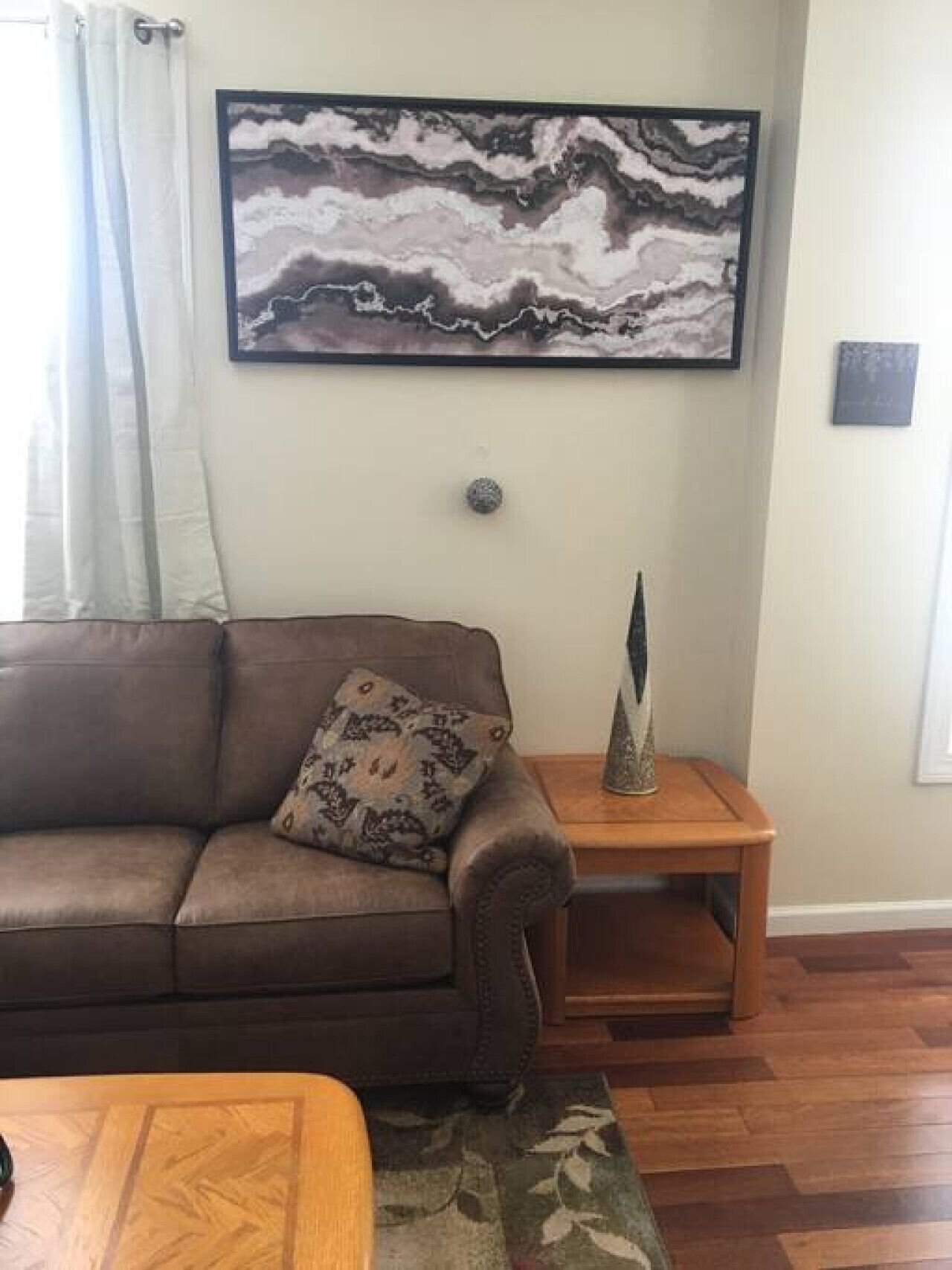Una sala de estar con un sofá marrón, un cojín estampado, una mesa de centro de madera, una obra de arte en mármol en la pared, una pequeña escultura decorativa y una mesita de noche de madera.