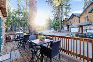Une terrasse en bois avec des sièges extérieurs, entourée de grands arbres et offrant une vue sur une rue.