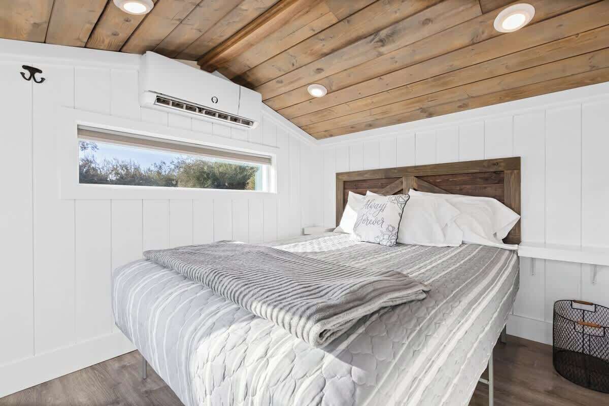 Ein Schlafzimmer mit Holzbalkendecke, weißen Wänden, einem großen Bett mit grau-weiß gestreifter Bettdecke und einem Fenster mit Blick auf Bäume.