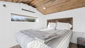 Une chambre avec un plafond en bois, des murs blancs, un grand lit recouvert d’une couette à rayures grises et blanches, et une fenêtre donnant sur des arbres.