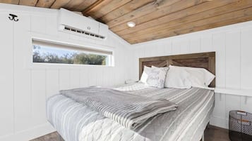 Ein Schlafzimmer mit Holzbalkendecke, weißen Wänden, einem großen Bett mit grau-weiß gestreifter Bettdecke und einem Fenster mit Blick auf Bäume.