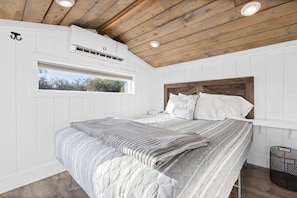 A bedroom with a wooden ceiling, white walls, a large bed with a grey and white striped bedspread, and a window with a view of trees.