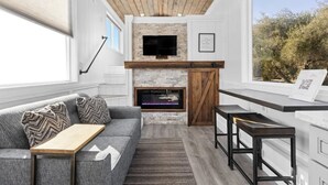 A modern living room with a fireplace, a sofa, a coffee table, and a TV mounted on the wall. - Charming Tiny Home in Olive Grove - Near Sequoias (Lindsay)