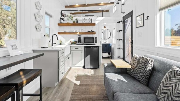 A compact kitchen with white cabinetry, a built-in microwave, and a small refrigerator. There is a wooden bench at the kitchen counter and a gray sofa with patterned cushions in the living area. - Charming Tiny Home in Olive Grove - Near Sequoias (Lindsay)