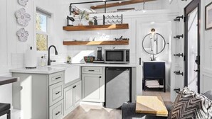 A compact kitchen with white cabinetry, a built-in microwave, and a coffee machine. There is a wooden shelf above the sink and a small refrigerator below. - Charming Tiny Home in Olive Grove - Near Sequoias (Lindsay)