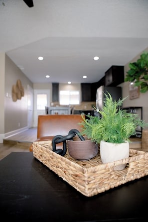 A modern living room with a black table, a woven tray with a potted plant and a black object, and a kitchen in the background.