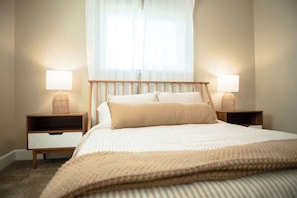 A neatly made bed with a wooden headboard, two bedside tables with lamps, and a window with sheer curtains.