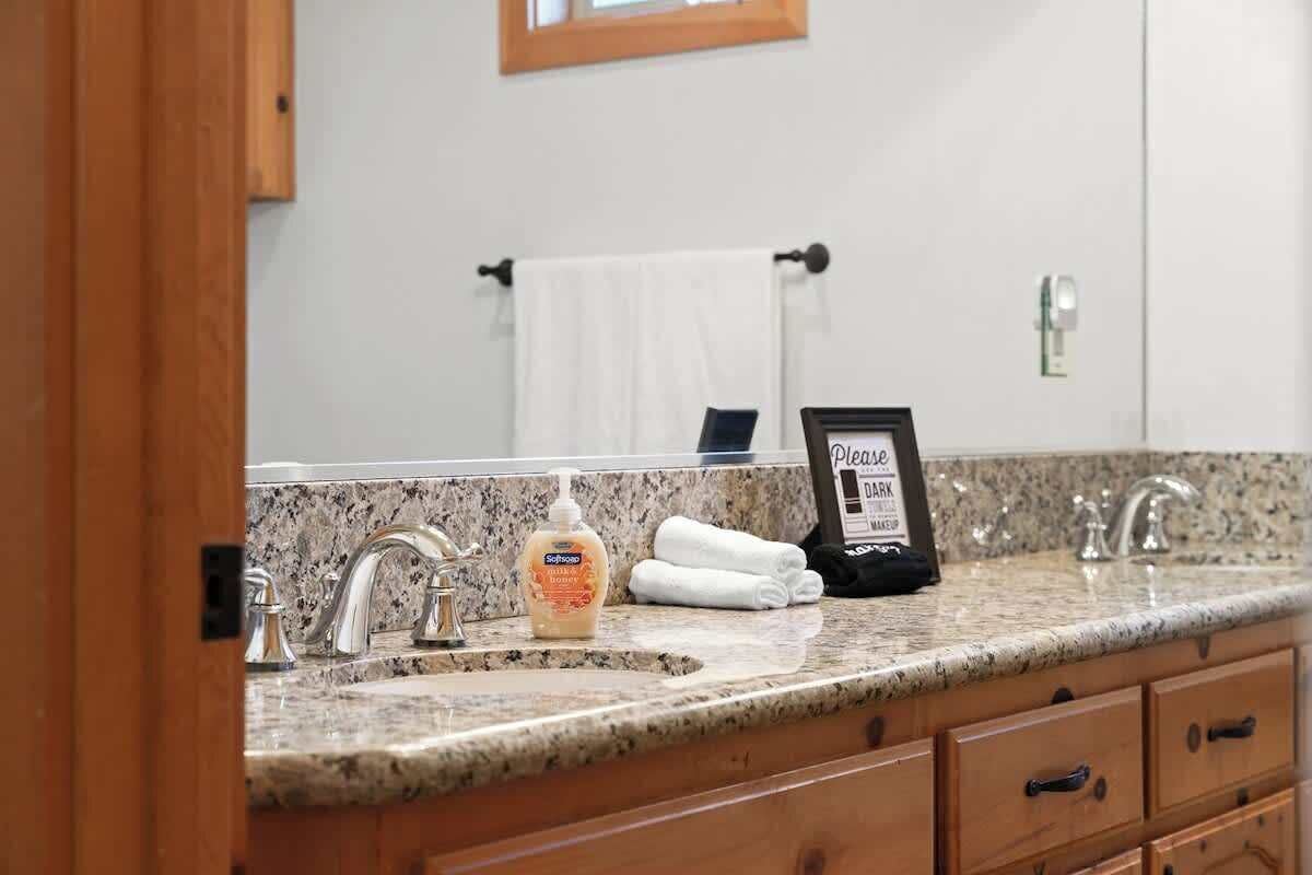A bathroom with a granite countertop, two sinks, and a mirror. There is a bottle of soap and a sign on the counter.