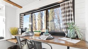 A dining area with a wooden table, two chairs, a window with curtains, and a view of trees.