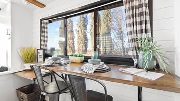 A dining area with a wooden table, two chairs, a window with curtains, and a view of trees.
